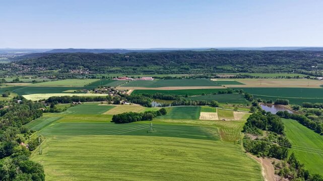 Elevated rural panorama showing green meadow with mowing lines, rectangular crop parcels, reflective ponds, forested ridge, hedgerows, and scattered farm buildings across tranquil countryside