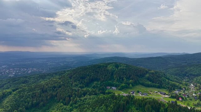 Elevated oblique aerial view of rolling forested hills, scattered rural houses, and winding roads beneath expansive cloud cover, where openings gradually reveal sunbeams and brighter light across