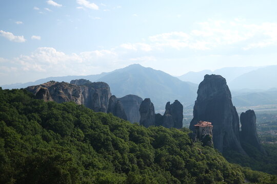 Meteora monastery perched on a rock formation above lush forest with a pointed mountain peak in the distance, Greece