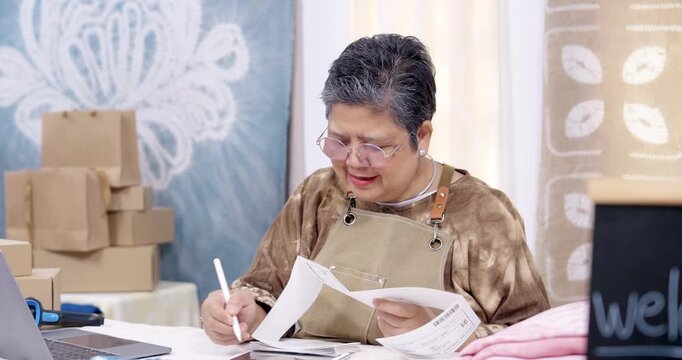 Senior asian seller smiles, flips invoice pages, writes order notes, checks barcode label on paperwork, prepares parcel box stack for small business shipping, home studio workshop routine continues.