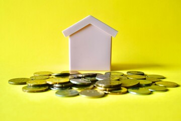 Centered shot of a toy house model sitting behind a scattered pile of Malaysian coins on a plain yellow background.  © Akram