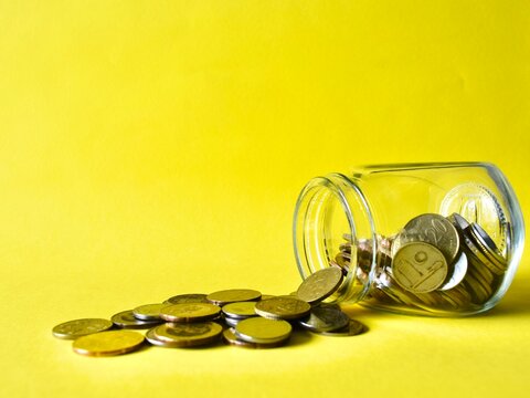 An overturned clear glass jar with Malaysian ringgit coins spilling out onto a vibrant yellow background, leaving ample copy space on the left.
