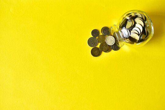 Minimalist overhead flat lay of a clear glass jar tipped on its side, spilling a neat cluster of metallic coins onto a bright yellow background with extensive negative space.