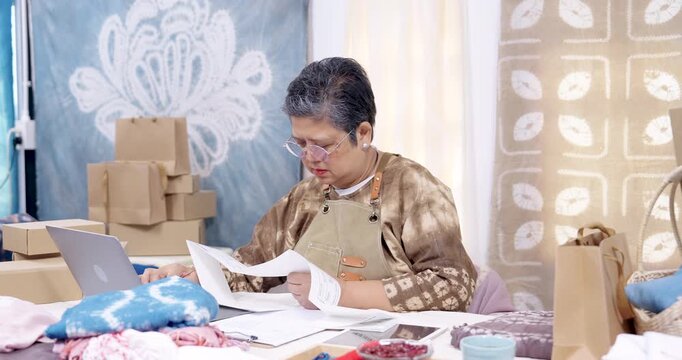 Senior asian retired businesswoman reviews invoice paper, checks online order on laptop, confirms packing list, organizes tie-dye shibori fabric, ready for parcel shipping workflow from home studio.