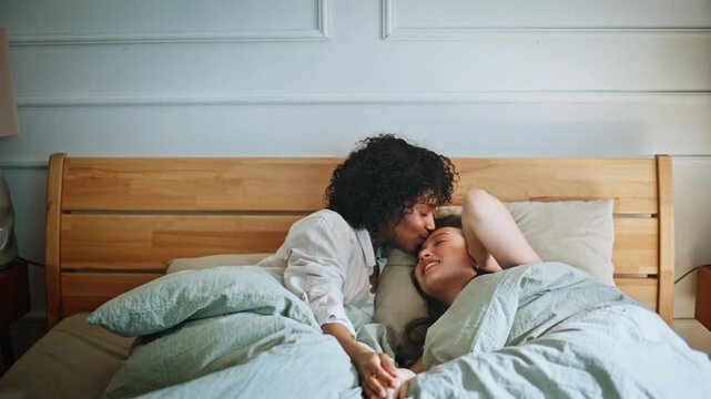 Lesbian couple cuddling bed morning. Two women holding hands, forehead kiss.