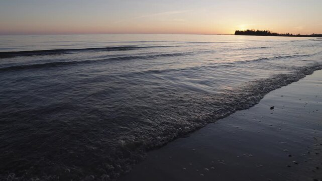 Soft waves gently wash over the flat sandy shoreline during a spring sunset on the Bruce Peninsula. The calm water of Lake Huron reflects the warm twilight sky on a quiet evening in Ontario.
