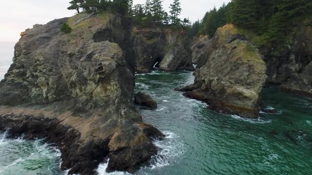 A long cinematic wide-angle aerial drone shot through the rugged cliffs, unique rock formations, sea arches, and coastal forest at Samuel H. Boardman State Scenic Corridor on Oregon's southern coast.