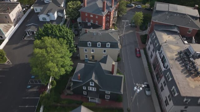 Pale yellow Victorian house with steep gable roofs stands among historic buildings in Salem Massachusetts, car travels quiet street past parking lots and colonial homes. Aerial drone flyover shot