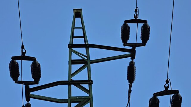 Bright daylight reveals green lattice transmission tower with suspended insulators and thick overhead cables, creating strong geometric lines against clear open blue sky, with power, electricity