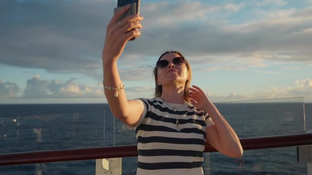 Young woman in striped shirt takes selfies with smartphone on cruise liner deck in Caribbean Sea. She poses playfully, touching hair and blowing kisses against ocean horizon and dramatic cloudy sky.
