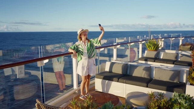 Happy woman in tropical shirt and sun hat takes selfie on luxury cruise liner deck in Caribbean Sea. Glass railings frame ocean horizon with elegant lounge seating and succulent plants on sunny day.