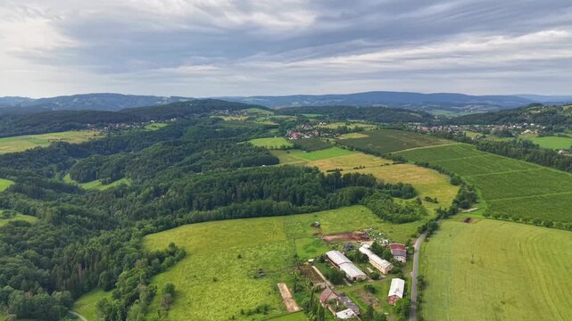 Wide aerial view reveals patchwork fields, hedgerows, orchards, farm buildings, winding roads, and distant villages across rolling countryside with muted light and calm rural atmosphere