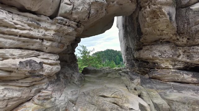 Natural arch opening in rocky canyon reveals distant forest and soft daylight, with layered limestone and sandstone surfaces, rugged textures, cliffside detail, and calm scenic travel atmosphere
