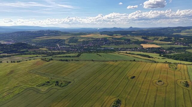 Wide rural landscape from above shows patchwork farmland, curved crop rows, circular field patterns, scattered farmhouses, distant village, gentle hills, green pastures, and clear bright sky