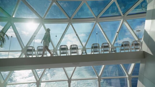Woman wearing hat and tropical print walks across elevated glass bridge on luxury cruise ship with stunning geometric dome ceiling, blue sky with white clouds.