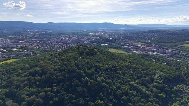 High view shows valley city bordered by forested hills, with trees in foreground, streets and parks below, low to mid rise buildings across urban spread, and wind turbines on distant ridge under