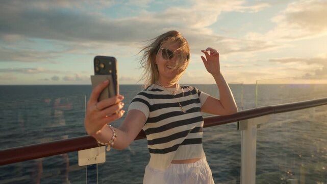 Young woman wearing striped shirt and sunglasses takes selfie photos with smartphone on cruise ship deck during golden hour sunset, ocean waves and dramatic cloudy sky visible in warm evening light.