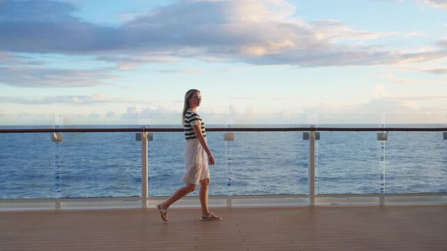 Young woman wearing striped nautical shirt and white shorts strolls leisurely across wooden cruise ship deck at sunset.