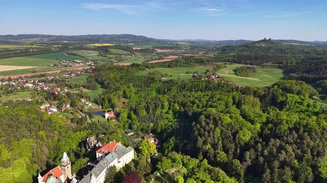 High altitude view reveals patchwork farmland, scattered villages, forest belts, rolling hills, and distant castle ruin beneath bright clear sky, creating spacious rural scenery with heritage