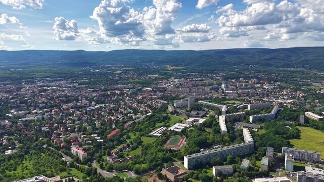 High altitude cityscape shows dense residential district set among rolling green hills, with towers, parks, roads, rooftops, sunlight, cloud cover, and distant mountain backdrop, with aerial, city