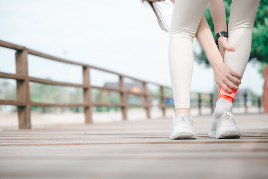 Woman suffers Achilles tendon ankle injury holding foot during workout, female athlete feels sudden sharp pain and joint inflammation while training on wooden boardwalk path in park