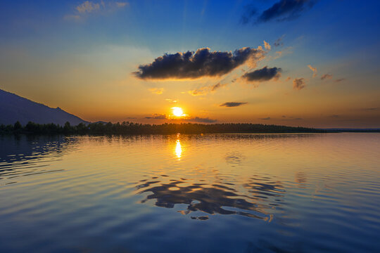 Beautiful sunset over Borovoe lake in Kazakhstan. Blue orange sky, bright sun golden reflections and clouds reflects on water. Dark silhouettes of mountains and forest away. Dusk Nature landscape.