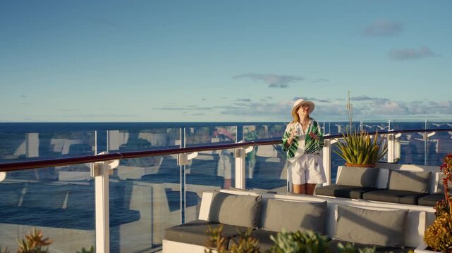 Stylish woman in white hat and tropical print shirt stands on luxury cruise liner deck sipping coffee while gazing at the calm Caribbean Sea waters under beautiful blue sky with scattered clouds.
