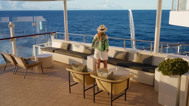 Woman in straw hat and tropical blouse stands on elegant cruise liner deck gazing at the Caribbean Sea. Luxury outdoor lounge with wicker chairs and cushioned seating overlooks deep blue ocean waters.