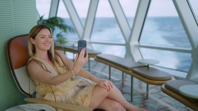 Young woman in yellow dress uses smartphone for video call while sitting in modern lounge chair aboard cruise liner in the Caribbean Sea. Ocean waves visible through panoramic windows.