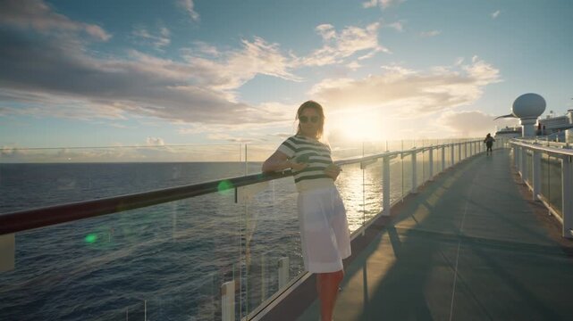 Young woman in striped shirt stands on cruise liner deck browsing smartphone during sunset in Caribbean Sea. Glass railings and ocean horizon create serene maritime travel atmosphere.