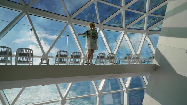 Stylish woman in sun hat and tropical print cover-up walks across modern glass skywalk aboard luxury cruise ship with geometric triangular ceiling architecture and panoramic ocean views on sunny day.
