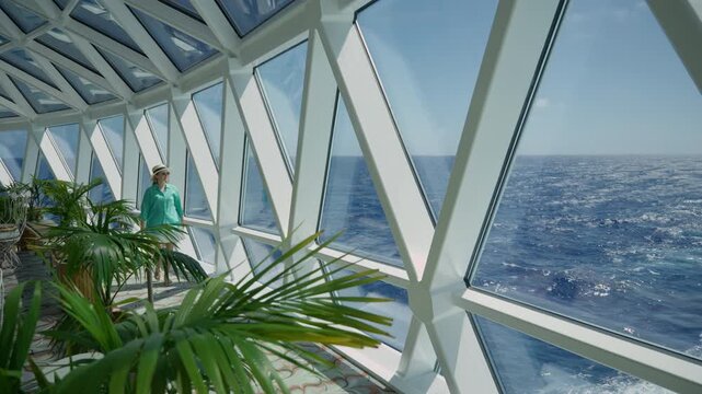 Smiling woman wearing turquoise shirt and straw hat walks through stunning geometric glass observation lounge on luxury cruise ship with panoramic ocean views and tropical palm plants in foreground.