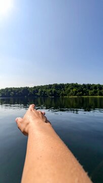 Person Reaching Hand Toward Calm Lake Water and Distant Forest Horizon Under Clear Blue Summer Sky