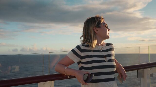 Young woman in striped shirt stands on cruise liner deck in Caribbean Sea during golden hour sunset. Wind blows through her hair as she relaxes with ocean views and dramatic clouds in the background.