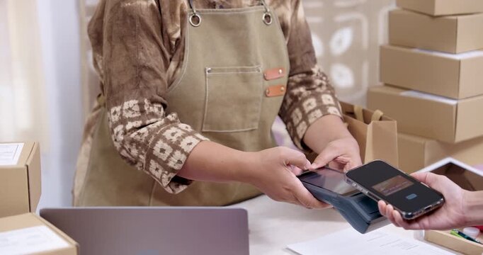 Slow motion wide shot, senior asian businesswoman accepts tap to pay phone transaction on POS terminal, checks paperwork order processing near laptop, boxes stacked, shibori workshop ships parcel.