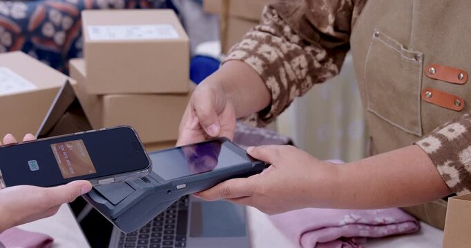 Retired asian woman entrepreneur angles EDC machine toward customer smartphone, confirmation check appears, boxes stacked by laptop, cashless transaction supports handmade shibori textile shipment.