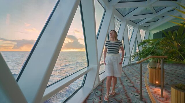 Young woman in striped shirt walks along glass-walled corridor on cruise liner in Caribbean Sea during golden hour. Panoramic ocean views through geometric windows with sunset sky and palm plants.