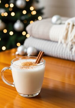 A glass mug of creamy eggnog with cinnamon sticks sits on a wooden table near a glowing Christmas tree and soft blankets in a cozy home, festive, cream, warm