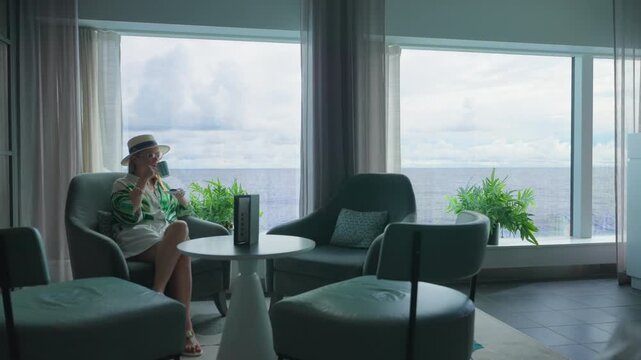 Stylish woman wearing straw hat and green striped shirt enjoys hot beverage in elegant cruise ship lounge with teal armchairs, white curtains, and stunning ocean view under cloudy tropical skies.