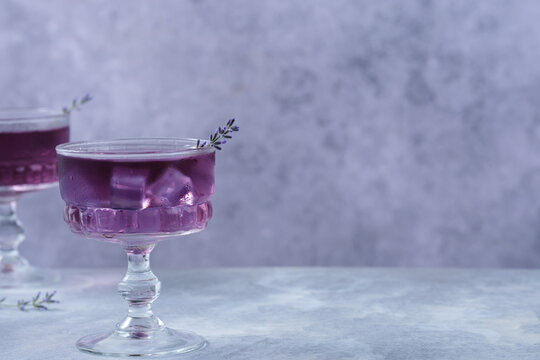 Refreshing purple lavender cocktail served in an elegant vintage coupe glass with ice cubes and a fresh lavender sprig. Minimalistic composition on gray background