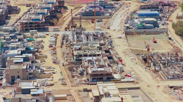 Expansive aerial perspective of a massive construction site, showcasing multiple buildings in various stages of completion, with heavy machinery and tower cranes actively developing the new area