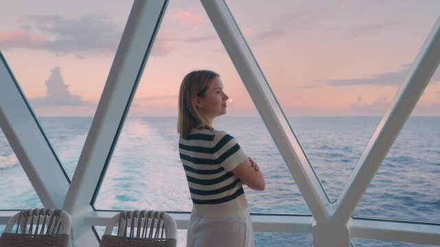 Young woman in striped shirt stands on modern cruise liner deck gazing at pink sunset over the Caribbean Sea.