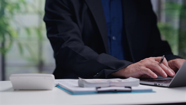 Male accountant wearing dark suit operates laptop keyboard, managing corporate financial budget, calculating taxes, counting cash money stack, planning business investment inside office.