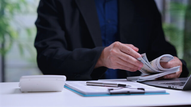 Businessman counts paper currency banknotes sitting inside office workspace near laptop computer calculator clipboard managing corporate budget financial audit revenue investment profit.