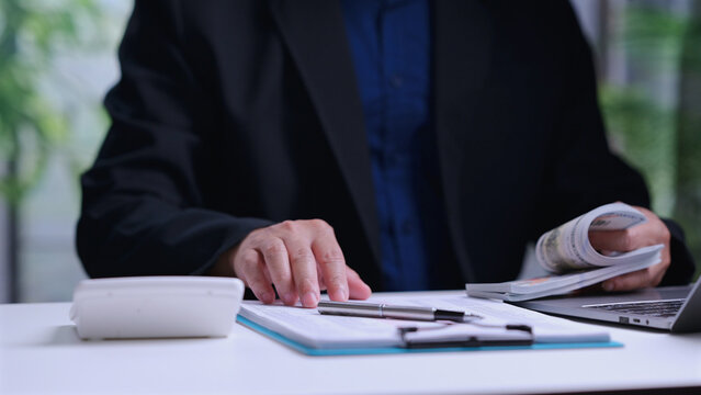 Male accountant wearing dark suit counts cash money banknotes sitting behind desk near laptop calculator clipboard document pen, managing financial budget, calculating business profit, planning.