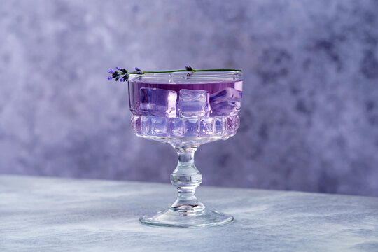 A glass filled with a purple drink sits on a gray background. Ice cubes fill the glass, and a sprig of lavender rests on the rim. 