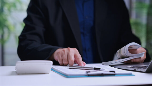 Businessman counts cash currency banknotes, analyzing financial document report, calculating budget spreadsheet inside office workspace, holding dollar bills near laptop computer keyboard.