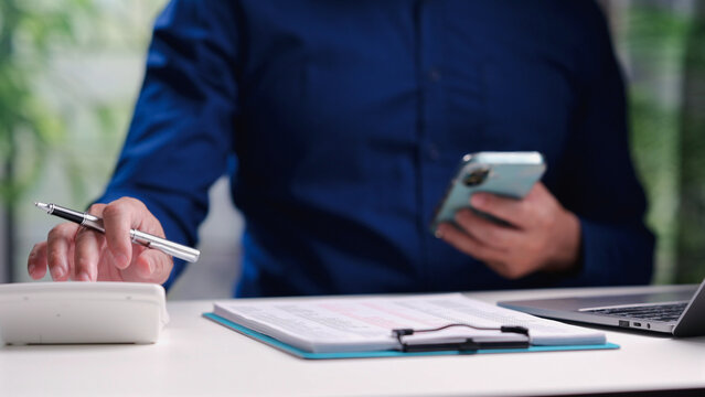 Male accountant holding smartphone calculates financial figures on clipboard document near laptop computer, managing corporate budget, tax planning, business investment paperwork inside office.