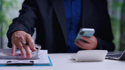 Fototapeta premium Corporate accountant holding mobile phone calculates financial budget touching cash paper document desk workspace laptop background investment planning tax analysis money management.