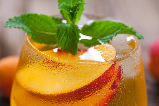 A glass filled with a beverage made of peach slices and mint leaves sits on a wooden table. The drink is cold and has condensation on the outside of the glass.